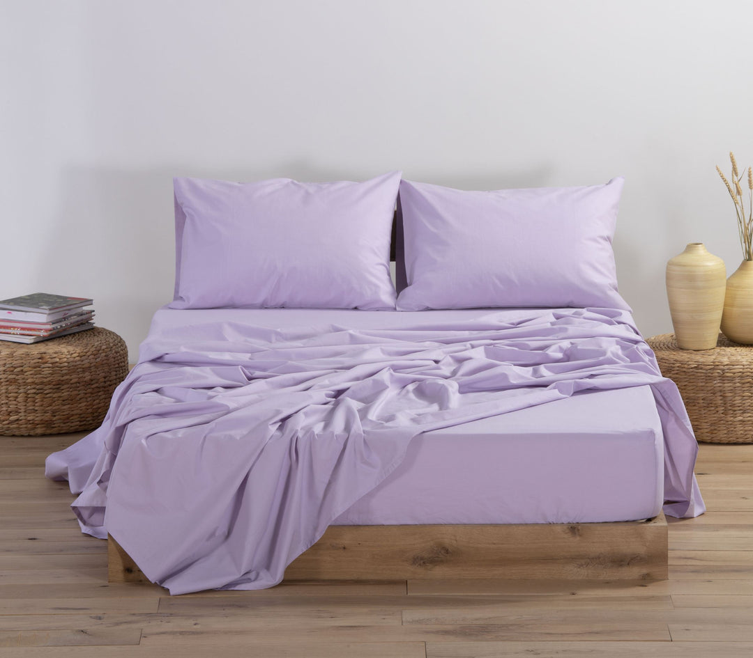 Σετ σεντονιών σε απαλή λιλά απόχρωση (lavender), στρωμένο σε ξύλινο διπλό κρεβάτι με ελαφρώς τσαλακωμένο πανωσέντονο. Το σετ περιλαμβάνει δύο μαξιλαροθήκες και ταιριαστό κατωσέντονο. Το δωμάτιο είναι διακοσμημένο με φυσικά στοιχεία, όπως ψάθινα κομοδίνα, κεραμικά βάζα και δεσμίδες από ξερά στάχυα.