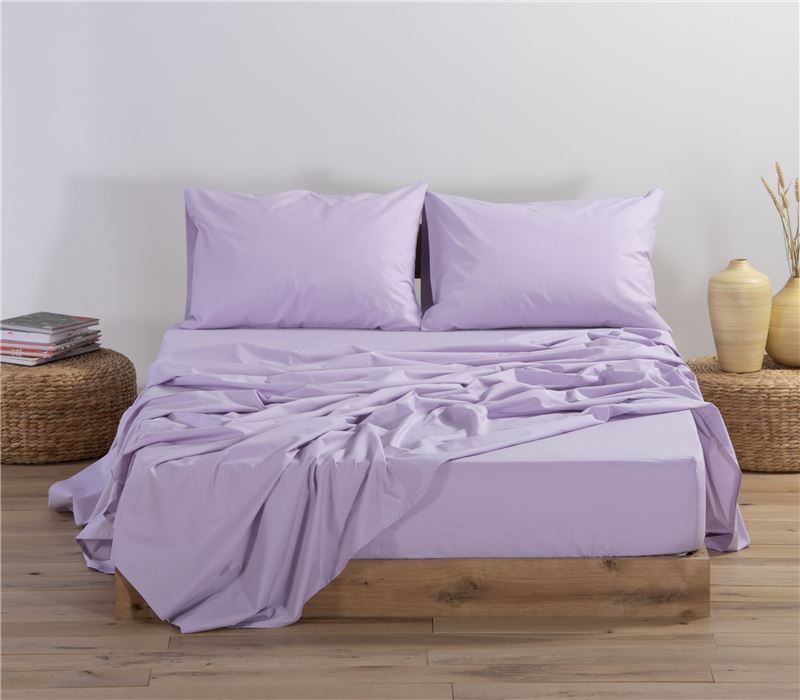 Σετ σεντονιών σε απαλή λιλά απόχρωση (lavender), στρωμένο σε ξύλινο διπλό κρεβάτι με ελαφρώς τσαλακωμένο πανωσέντονο. Το σετ περιλαμβάνει δύο μαξιλαροθήκες και ταιριαστό κατωσέντονο. Το δωμάτιο είναι διακοσμημένο με φυσικά στοιχεία, όπως ψάθινα κομοδίνα, κεραμικά βάζα και δεσμίδες από ξερά στάχυα.