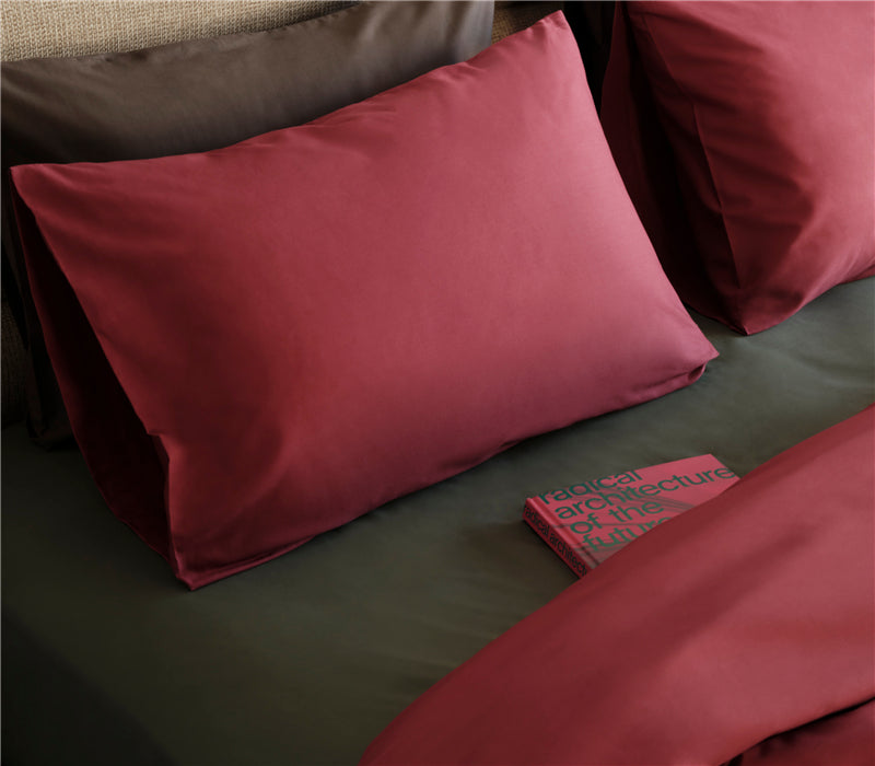 Μπορντό μαξιλαροθήκη τοποθετημένη σε σκούρο πράσινο κρεβάτι, με σκηνικό υπνοδωματίου και βιβλίο πάνω στα σεντόνια.