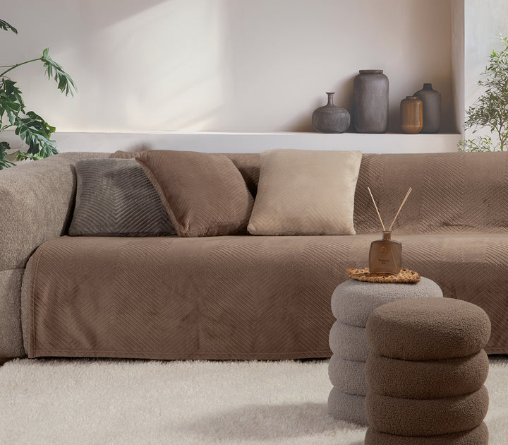 Καθιστικό με καναπέ σε μπεζ απόχρωση, διακοσμημένο με καφέ ριχτάρι βελουτέ και μαξιλάρια σε γήινους τόνους. Μοντέρνα διακόσμηση με απαλό χαλί και μινιμαλιστικά βάζα στο φόντο.