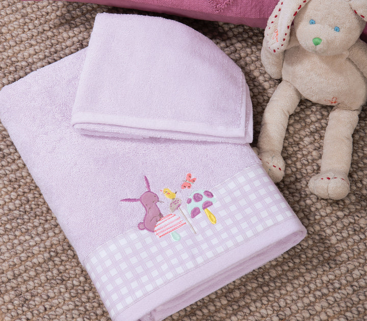Βρεφική πετσέτα σε λιλά χρώμα με κεντητό σχέδιο ζωάκια και μανιτάρια στη μπορντούρα, τοποθετημένη πάνω σε ψάθινο χαλί δίπλα σε λούτρινο κουνελάκι.