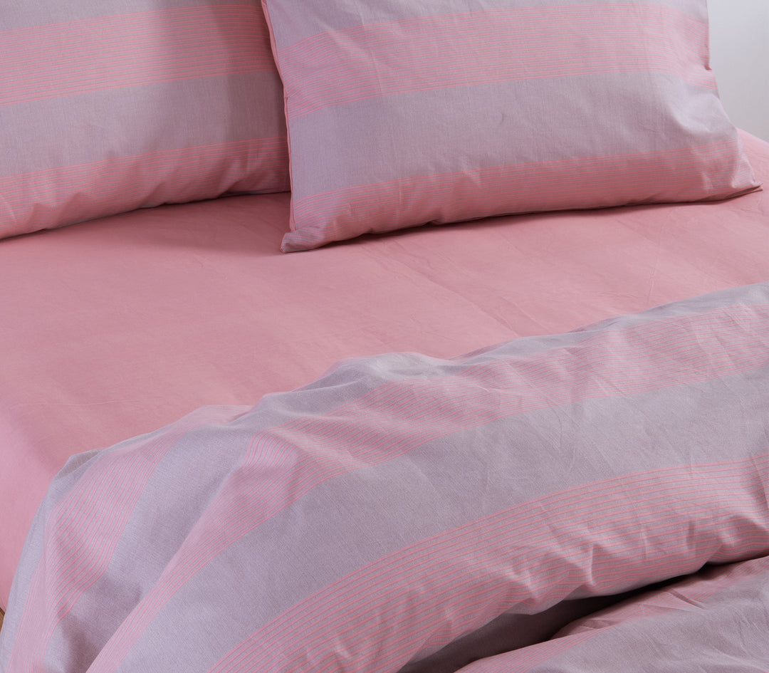 Κοντινή λήψη από ριγέ σεντόνια και μαξιλάρια σε ροζ και γκρι αποχρώσεις, στρωμένα σε κρεβάτι με ροζ κατωσέντονο.
