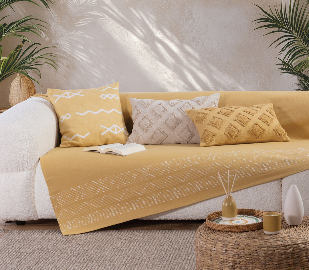 Ριχτάρι σε ζεστό μουσταρδί χρώμα με λευκά γεωμετρικά σχέδια, στρωμένο σε λευκό καναπέ σε φυσικό, φωτεινό περιβάλλον.