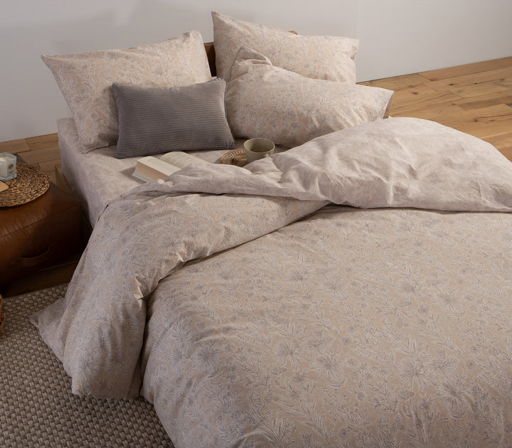 Υπνοδωμάτιο με μπεζ σεντόνια λουλουδάτο σχέδιο, διακοσμημένο με γκρι μαξιλάρι, βιβλίο και κούπα καφέ.