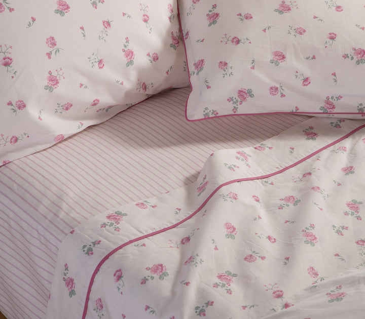 Κρεβάτι στρωμένο με σετ σεντονιών, λευκή βάση με ροζ λουλούδια και ριγέ κατωσέντονο.