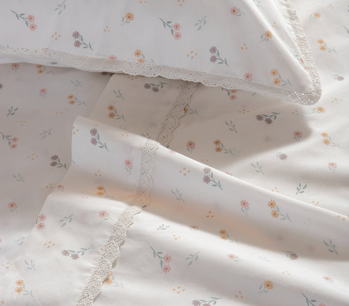 Κοντινή λήψη από σετ λευκών σεντονιών με μικρά λουλούδια σε ροζ και κίτρινους τόνους και δαντελένια διακόσμηση, που αποπνέουν κομψότητα και φρεσκάδα.