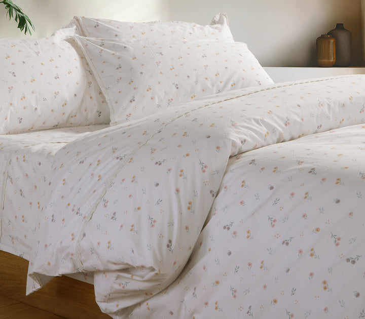 Διπλό κρεβάτι στρωμένο με σετ σεντόνια σε λευκή βάση με φλοράλ μοτίβο και δαντελένιες λεπτομέρειες, σε φωτεινό υπνοδωμάτιο με ξύλινη διακόσμηση.
