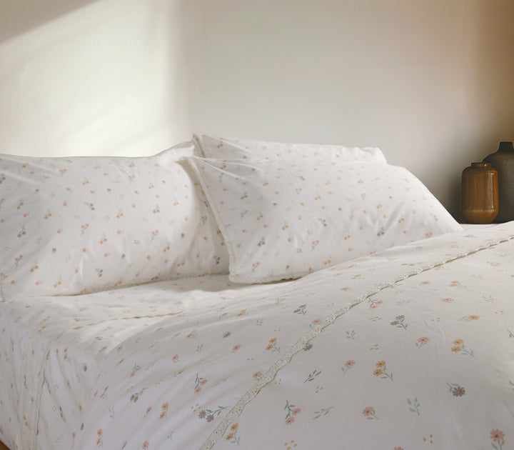 Κρεβάτι στρωμένο με σετ σεντονιών σε λευκή βάση με μικρά λουλούδια και λεπτομέρεια δαντέλας, σε φωτεινό δωμάτιο.