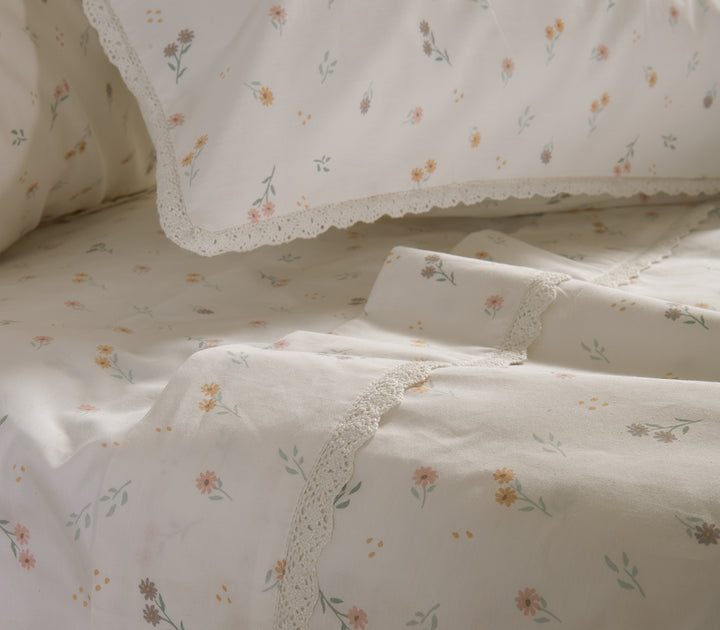 Λεπτομέρεια κρεβατιού στρωμένου με λευκά σεντόνια με μικροσκοπικά λουλούδια και δαντέλα στο τελείωμα, που δημιουργούν ρομαντική αίσθηση.