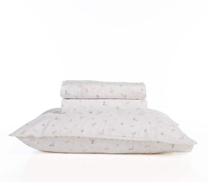 Σετ σεντόνια λευκής βάσης με μικρά λουλουδάκια σε παστέλ τόνους και δαντελένιο τελείωμα, τακτοποιημένα διπλωμένα πάνω σε μαξιλάρι σε λευκό φόντο.