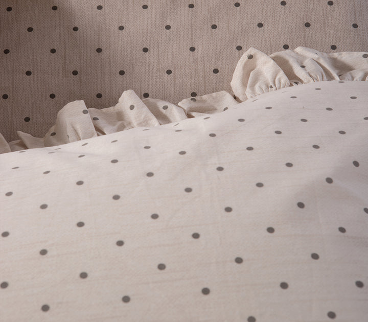 Κοντινή λήψη σε κρεβάτι στρωμένο με μπεζ σεντόνια πουά και μαξιλαροθήκες με βολάν, συνδυασμένα με μπεζ διακοσμητικό μαξιλάρι βελούδου.