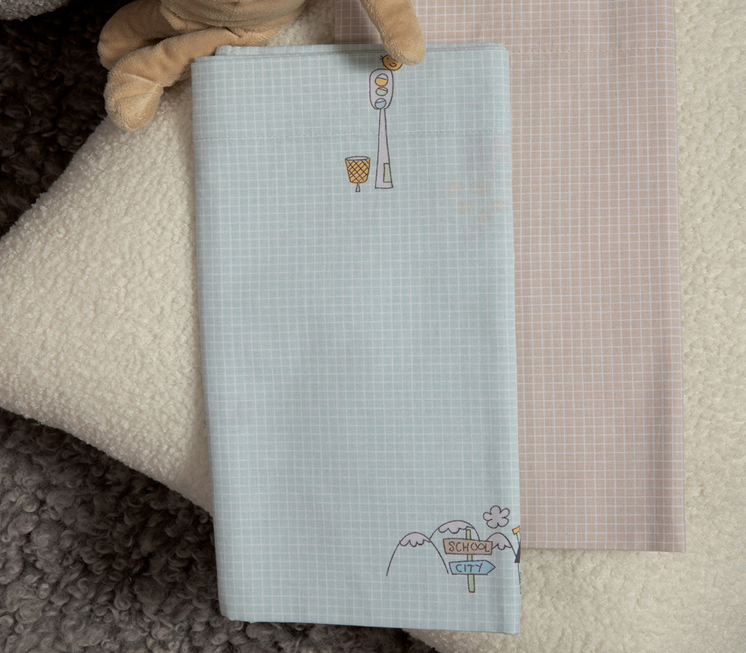 Βρεφικά σεντόνια διπλωμένα, σε γαλάζιο και μπεζ με καρό μοτίβο. Το γαλάζιο σεντόνι έχει χαριτωμένα σχέδια με φωτεινό σηματοδότη, καλαθάκι και πινακίδα που γράφει "School City", ενώ το μπεζ παραμένει μινιμαλιστικό με το ίδιο καρό μοτίβο. Φωτογραφημένα πάνω σε απαλή φλοκάτη, δίπλα σε λούτρινο παιχνίδι.