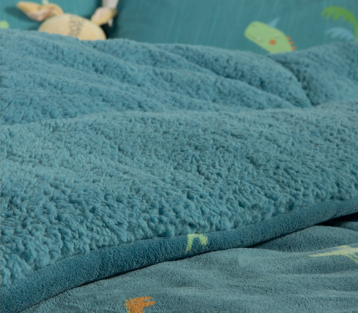 Κοντινή εικόνα παιδικού δωμάτιου για αγόρι με στρωμένο κρεβάτι σε μπλε και πετρόλ αποχρώσεις με κουβερτοπάπλωμα που έχει σχέδιο από δεινόσαυρους και αντίστοιχα μαξιλάρια ύπνου.