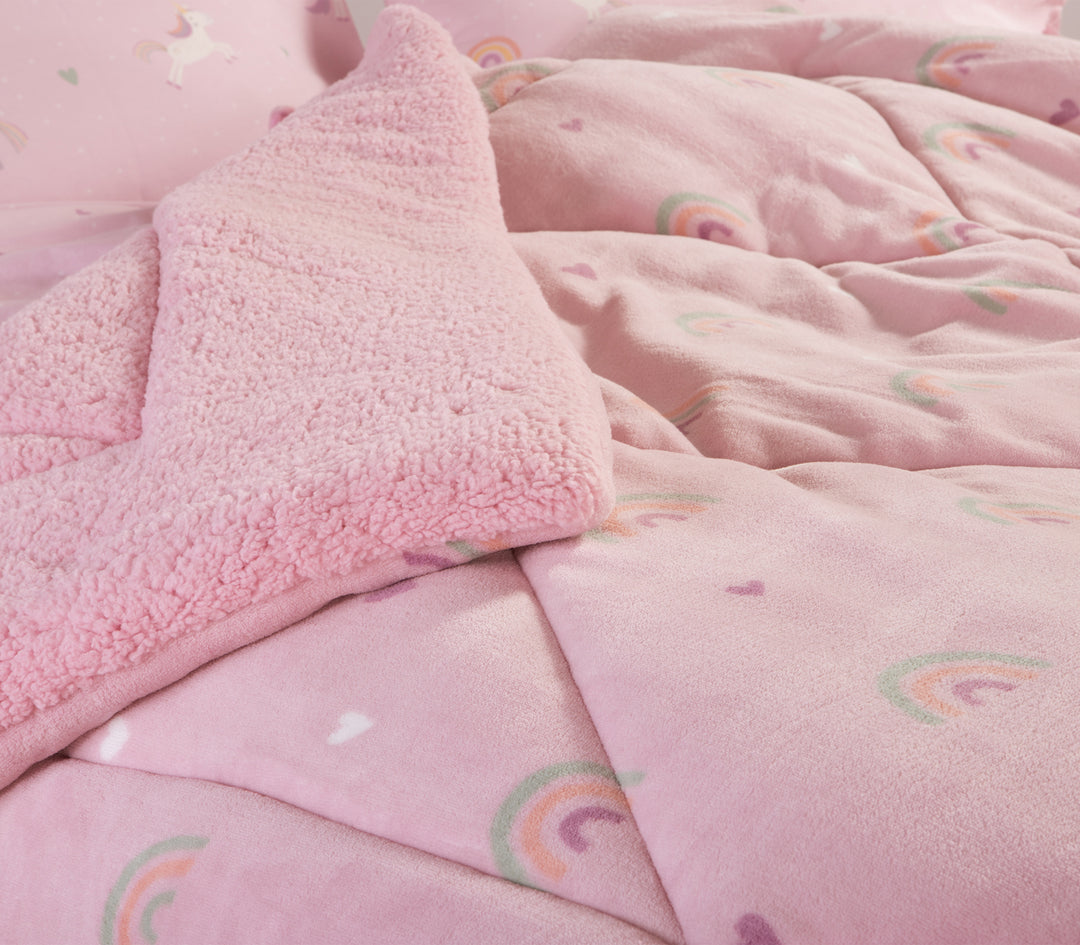 Κοντινή εικόνα παιδικού κρεβατιού για κορίτσι  που περιέχει ροζ κουβερτοπάπλωμα με σχέδια από ουράνιο τόξο και μαξιλαροθήκες με σχέδιο από μονοκέρους.