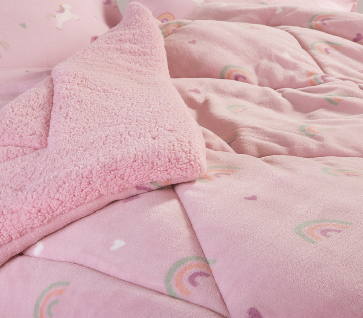 Κοντινή εικόνα παιδικού κρεβατιού για κορίτσι  που περιέχει ροζ κουβερτοπάπλωμα με σχέδια από ουράνιο τόξο και μαξιλαροθήκες με σχέδιο από μονοκέρους.