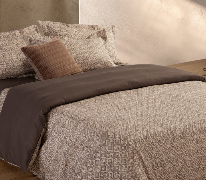 Κρεβάτι με σετ σεντόνια σε καφέ και μπεζ αποχρώσεις με γεωμετρικό μοτίβο και διακοσμητικό μαξιλάρι.