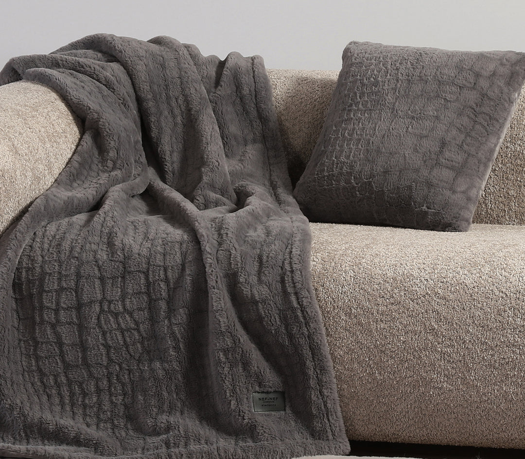 Γκρι κουβέρτα με μαξιλάρι σε ίδια υφή επάνω σε μπεζ καναπέ.