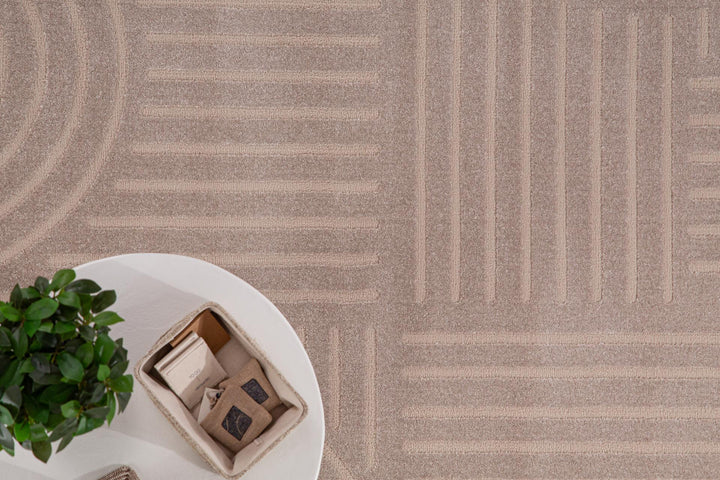 Μπεζ χαλί Organic Royal Carpet με γεωμετρικές γραμμές και καμπύλες, πλαισιωμένο από λευκό στρογγυλό τραπέζι με φυτό και κουτί οργάνωσης.