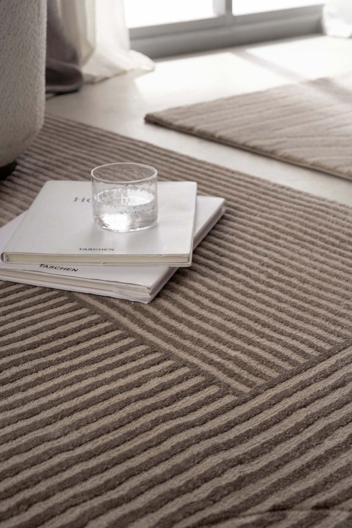 Μοντέρνο χαλί Organic Royal Carpet σε μπεζ χρώμα με γεωμετρικό σχέδιο, κοντινή λεπτομέρεια με βιβλία και γυάλινο ποτήρι στο πάτωμα.