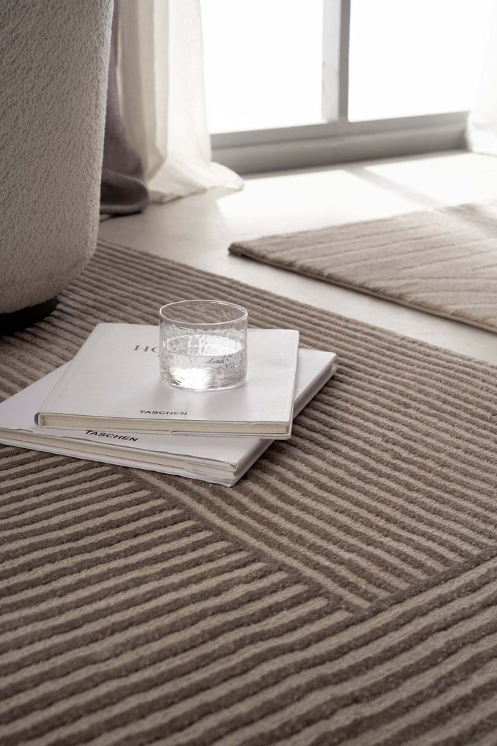 Κοντινή λήψη χαλιού Organic Royal Carpet σε μπεζ αποχρώσεις με γεωμετρικό σχέδιο, με βιβλία και ποτήρι πάνω στο πάτωμα.