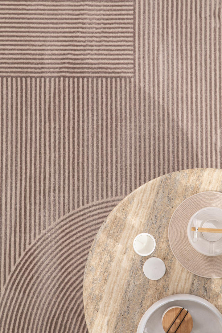 Μπεζ χαλί Organic Royal Carpet με γεωμετρικό σχέδιο από κυκλικές και ευθείες γραμμές, συνδυασμένο με στρογγυλό τραπέζι σε φυσική πέτρα.