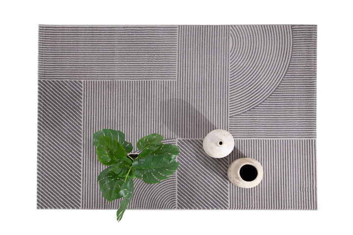 Χαλί Organic Royal Carpet σε γκρι και εκρού με μοντέρνο γεωμετρικό σχέδιο, διακοσμημένο με φυτά και κεραμικά βάζα σε λευκό φόντο.