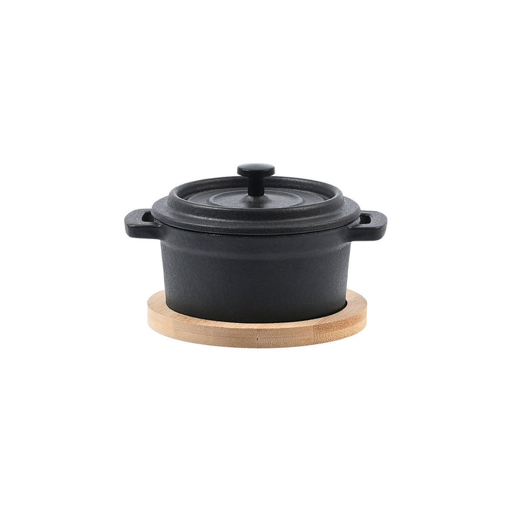 Μικρή μαύρη μαντεμένια κατσαρόλα με καπάκι, στηριγμένη σε στρογγυλή βάση από bamboo, με καθαρή και ισορροπημένη παρουσίαση.