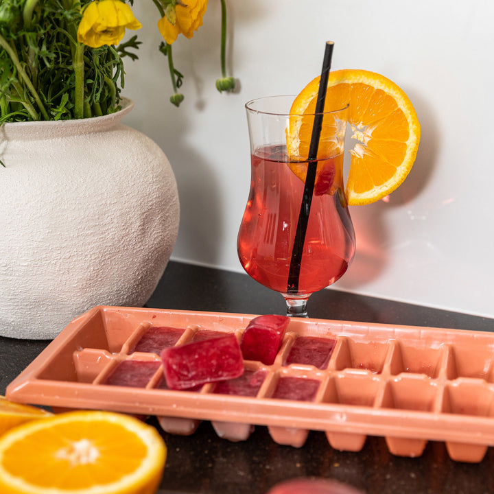 Η παγοθήκη Estia με κόκκινα παγάκια δίπλα σε ποτήρι με κοκτέιλ και φέτα πορτοκαλιού.