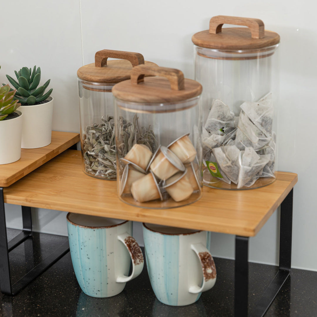 Γυάλινα δοχεία με ξύλινα καπάκια πάνω σε ξύλινο ράφι κουζίνας, γεμάτα με κάψουλες καφέ, αποξηραμένα βότανα και φακελάκια τσαγιού, δίπλα σε διακοσμητικά φυτά και κούπες.