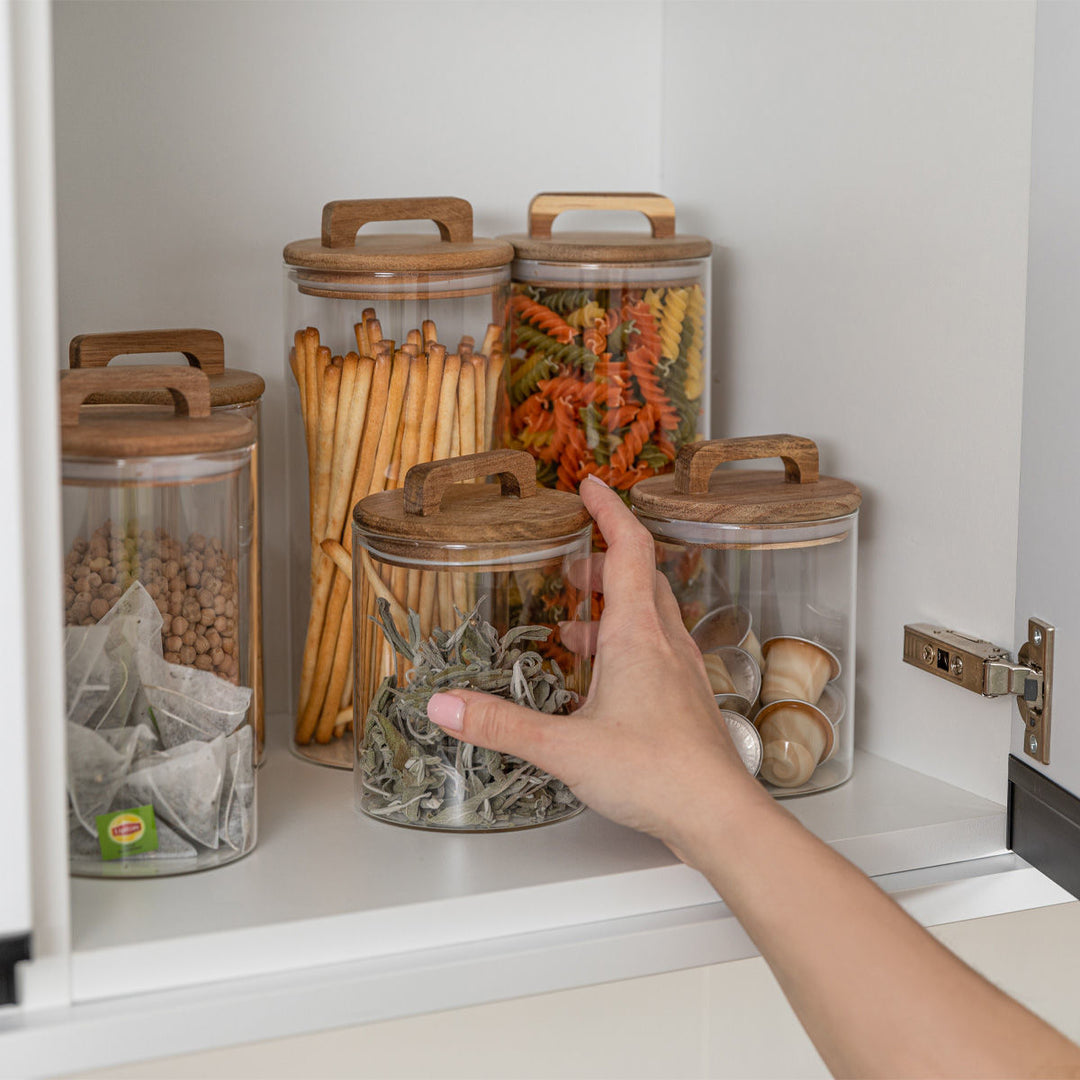 Γυάλινα δοχεία αποθήκευσης με ξύλινα καπάκια τοποθετημένα μέσα σε ντουλάπι κουζίνας, γεμάτα με ζυμαρικά, παξιμάδια, τσάι και κάψουλες καφέ, δημιουργώντας μια οργανωμένη και ζεστή εικόνα κουζίνας.