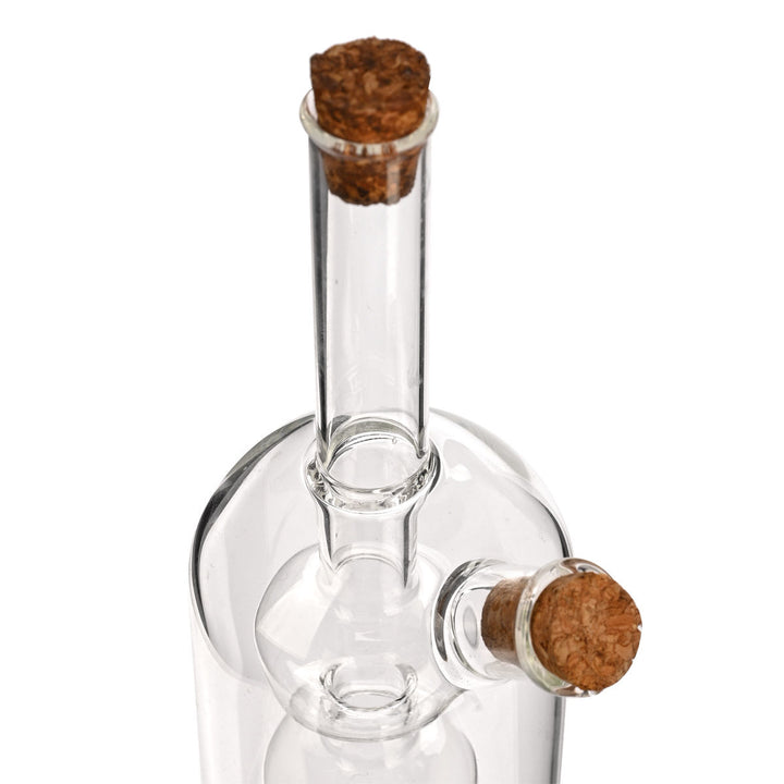 Κοντινή λήψη σε διάφανη γυάλινη φιάλη λαδιού με τριπλό εσωτερικό δοχείο και πώμα φελλού, σε λευκό φόντο.