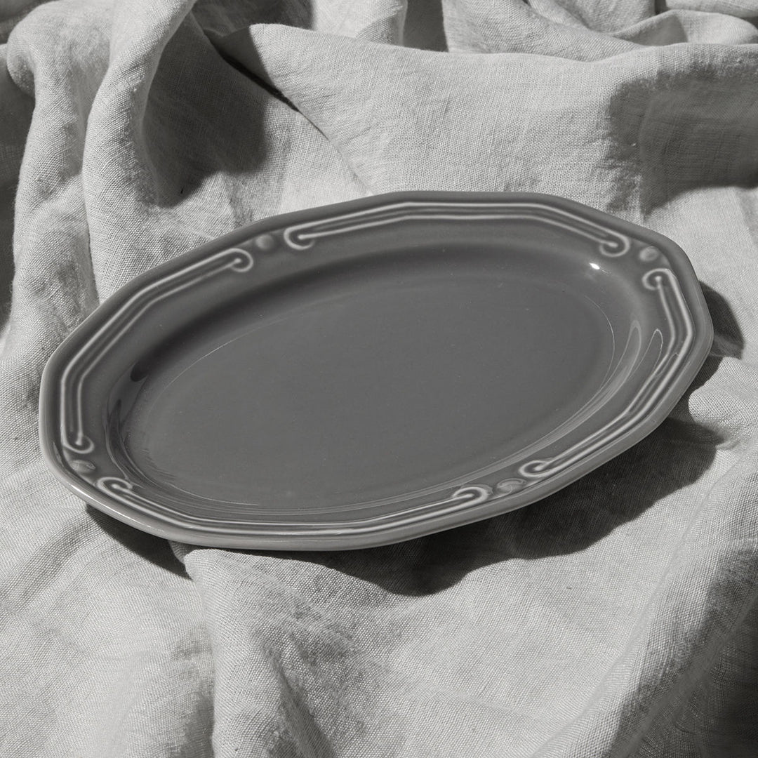 Γκρι οβάλ πιατέλα Athenee με ανάγλυφο διακοσμητικό τελείωμα, τοποθετημένη πάνω σε ανοιχτόχρωμο υφασμάτινο φόντο
