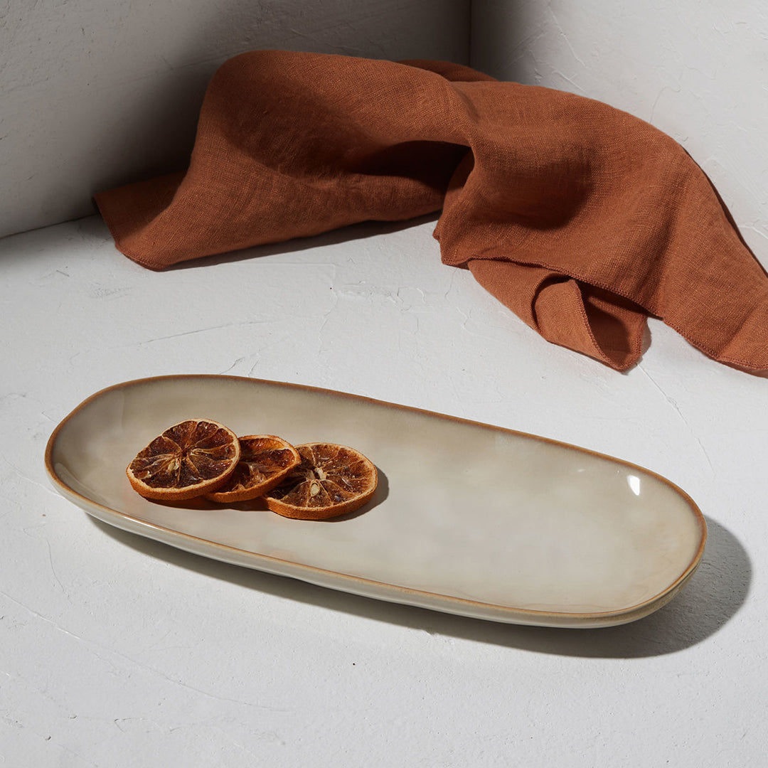 Οβάλ πιατέλα stoneware σε ivory απόχρωση με διακριτικό γυαλιστερό φινίρισμα, τοποθετημένη σε φωτεινό φόντο με υφασμάτινη λεπτομέρεια και αποξηραμένα εσπεριδοειδή.