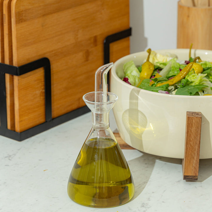 Γυάλινη κανάτα λαδιού με πράσινο ελαιόλαδο τοποθετημένη σε πάγκο κουζίνας, δίπλα σε σαλατιέρα και ξύλινα σκεύη.