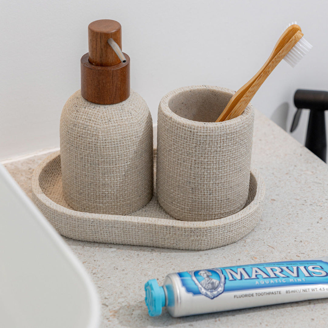 Μπεζ dispenser σαπουνιού Jute με ξύλινη αντλία και λεπτό μεταλλικό στόμιο, τοποθετημένο σε πάγκο μπάνιου μαζί με θήκη οδοντόβουρτσας στο ίδιο σχέδιο, δημιουργώντας φυσικό και ζεστό αποτέλεσμα.