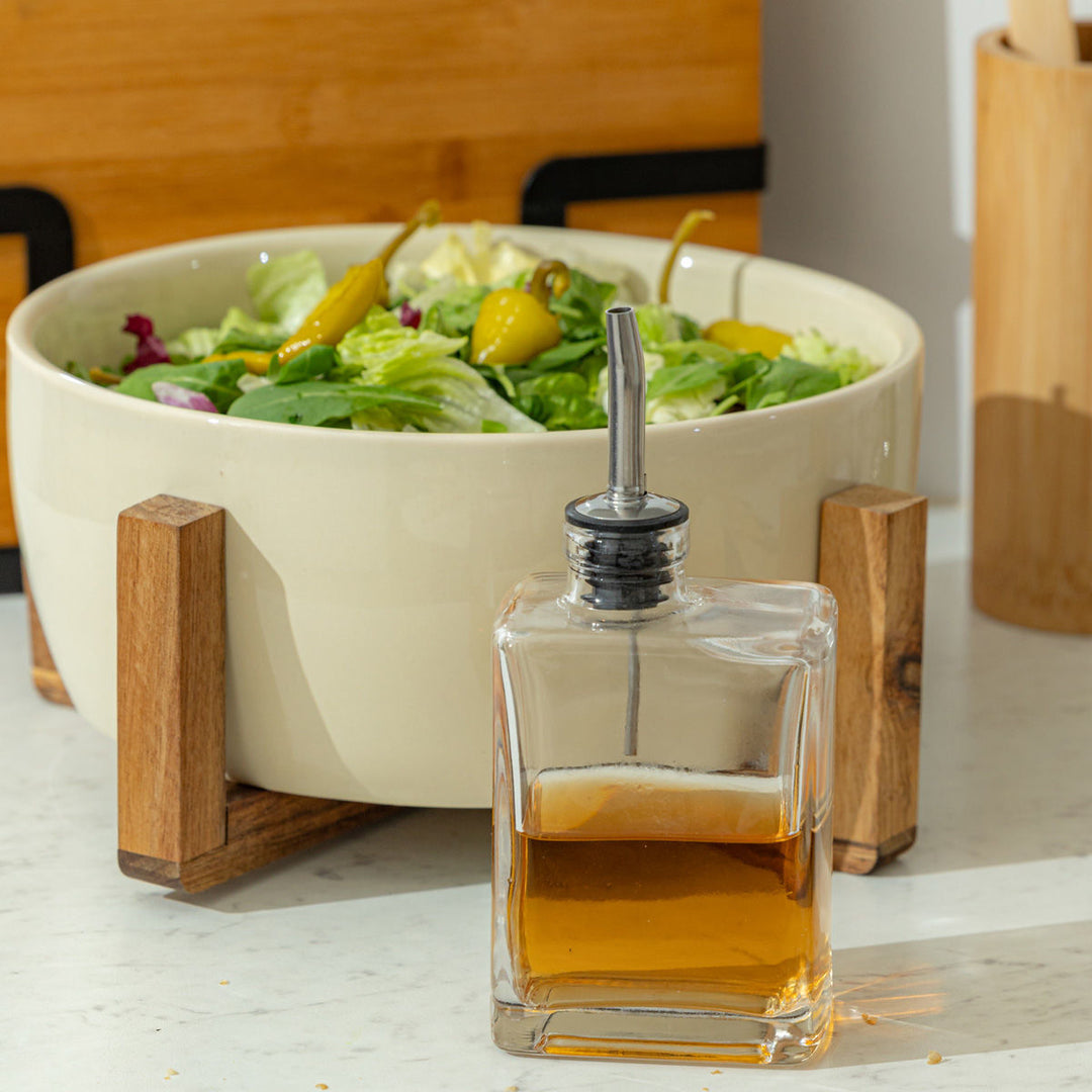Γυάλινο δοχείο λαδιού με ανοξείδωτο στόμιο, τοποθετημένο σε πάγκο κουζίνας μπροστά από μπολ σαλάτας με πράσινα φύλλα και πιπεριές, σε φωτεινό και φυσικό περιβάλλον κουζίνας.