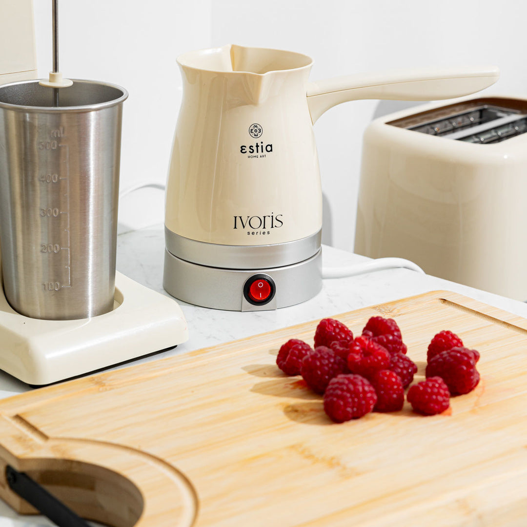 Ηλεκτρικό μπρίκι Estia Ivoris Series επάνω σε πάγκο κουζίνας, δίπλα σε ξύλο κοπής με φρέσκα βατόμουρα και συσκευές κουζίνας σε αντίστοιχες απαλές αποχρώσεις.