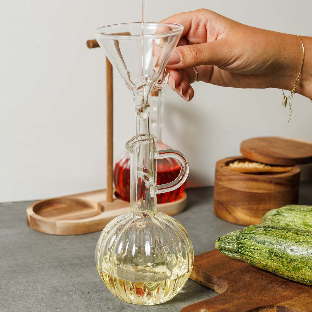 Γυάλινη φιάλη λαδιού με στρογγυλή βάση και λεπτό λαιμό, τοποθετημένη σε ξύλινη βάση, καθώς λάδι ρέει μέσα από γυάλινο χωνί. Στο φόντο διακρίνονται ξύλινα δοχεία κουζίνας και φρέσκα λαχανικά, δημιουργώντας φυσική, ζεστή ατμόσφαιρα κουζίνας.