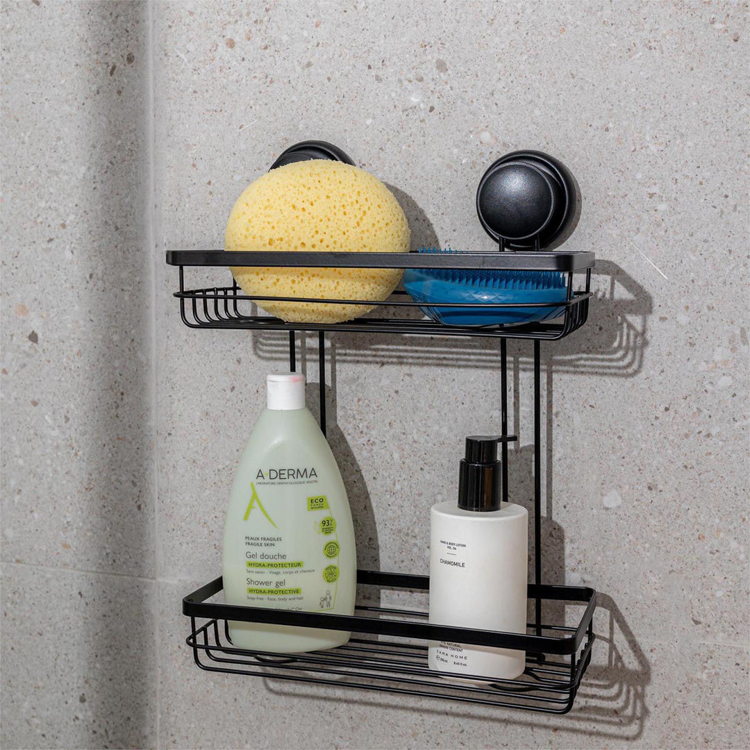 Μαύρη μεταλλική γωνιακή ραφιέρα μπάνιου με δύο επίπεδα, τοποθετημένη σε τοίχο με υφή πέτρας, γεμάτη με προϊόντα περιποίησης όπως σαμπουάν, αφρόλουτρο και σφουγγάρια μπάνιου.