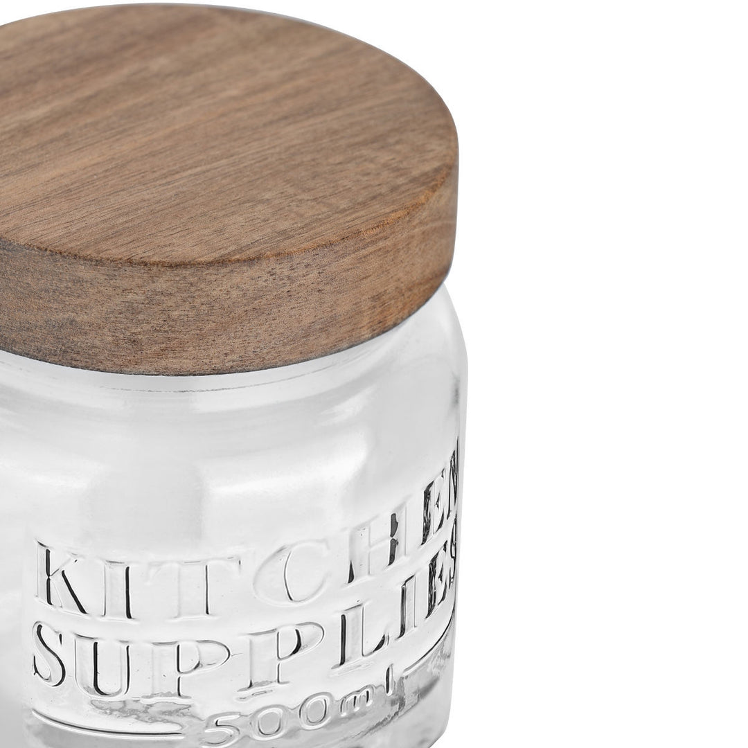 Κοντινή λήψη σε γυάλινο δοχείο αποθήκευσης Kitchen Supplies με ανάγλυφη επιγραφή, διαφανές σώμα και καπάκι από φυσικό ξύλο, ιδανικό για κουζίνα.