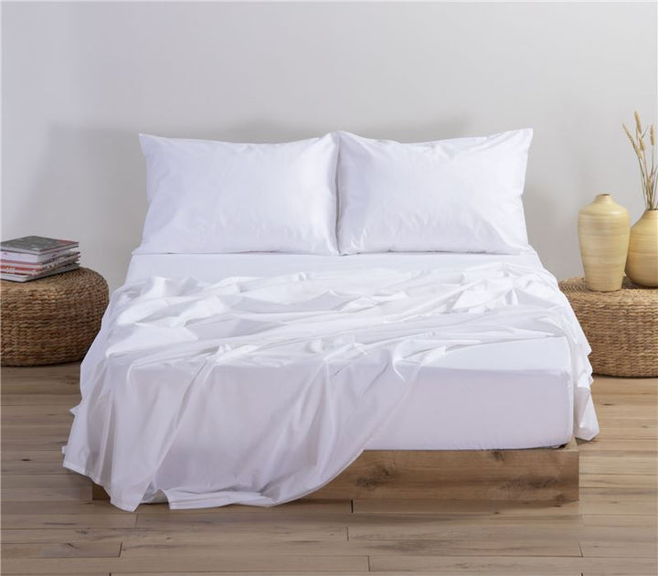 Κρεβάτι με μονόχρωμο σετ σεντονιών σε λευκή απόχρωση, που περιλαμβάνει πανωσέντονο, κατωσέντονο και δύο μαξιλαροθήκες, τοποθετημένο σε υπνοδωμάτιο με φυσική διακόσμηση και ξύλινο δάπεδο.