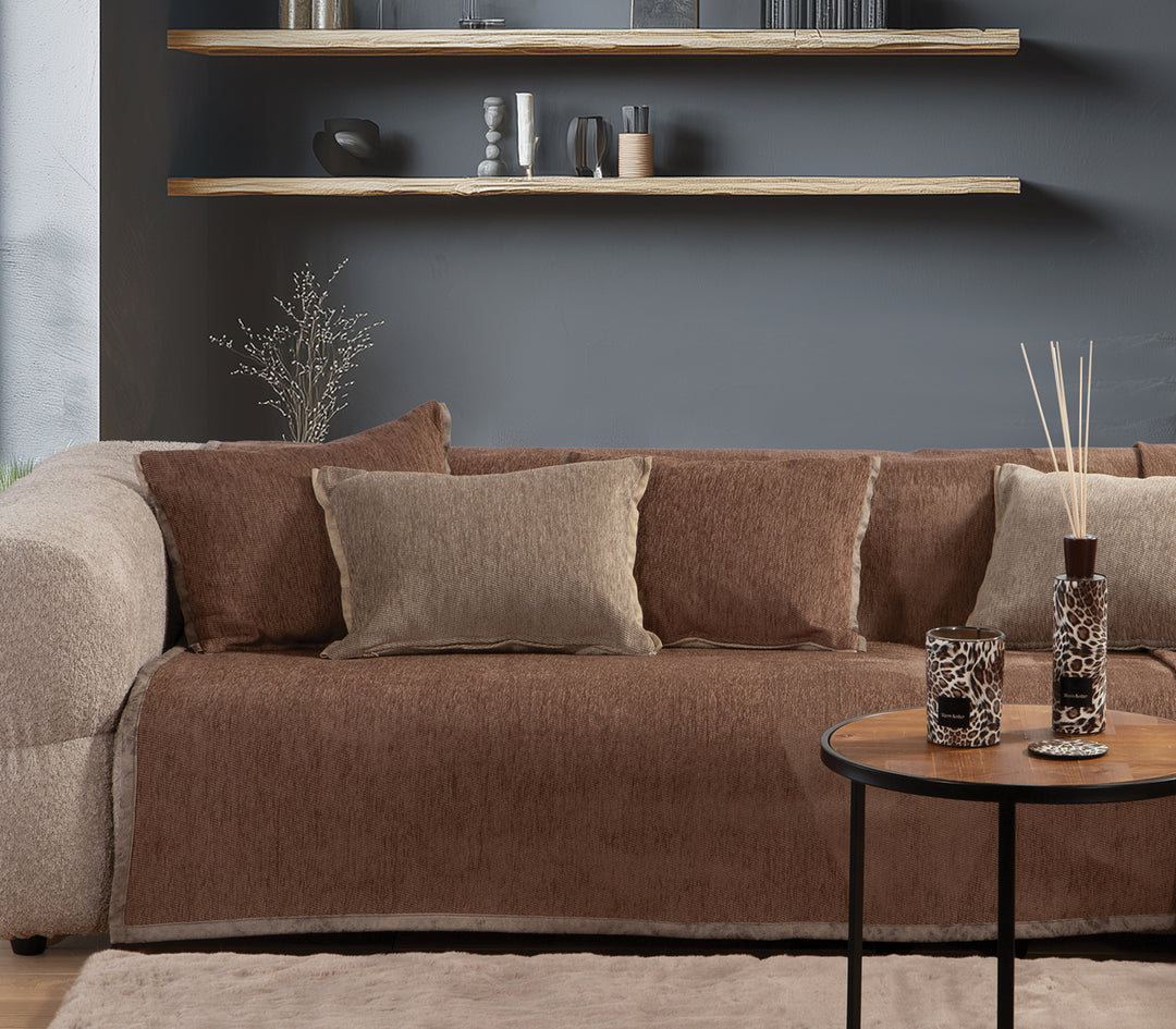 Καναπές διακοσμημένος με καφέ ριχτάρι και μαξιλάρια σε γήινες αποχρώσεις, σε μοντέρνο σαλόνι με γκρι τοίχο και ράφια.