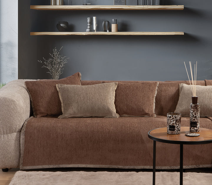 Καναπές διακοσμημένος με καφέ ριχτάρι και μαξιλάρια σε γήινες αποχρώσεις, σε μοντέρνο σαλόνι με γκρι τοίχο και ράφια.