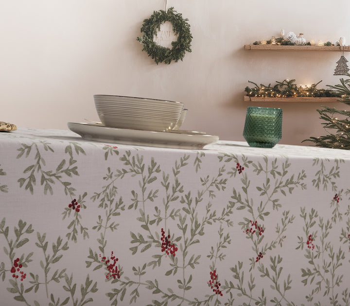 Χριστουγεννιάτικο τραπεζομάντηλο με σχέδιο από πράσινα κλαδιά και κόκκινους καρπούς, στρωμένο σε τραπέζι με πιάτα και πράσινο κερί, σε γιορτινό φόντο με στεφάνι και φωτισμένο δέντρο.
