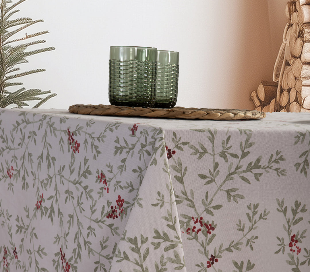 Χριστουγεννιάτικο τραπεζομάντηλο σε λευκό χρώμα με μοτίβο από πράσινα φύλλα και κόκκινους καρπούς, στρωμένο σε τραπέζι με ψάθινο σουπλά και γυάλινα ποτήρια σε πράσινη απόχρωση.