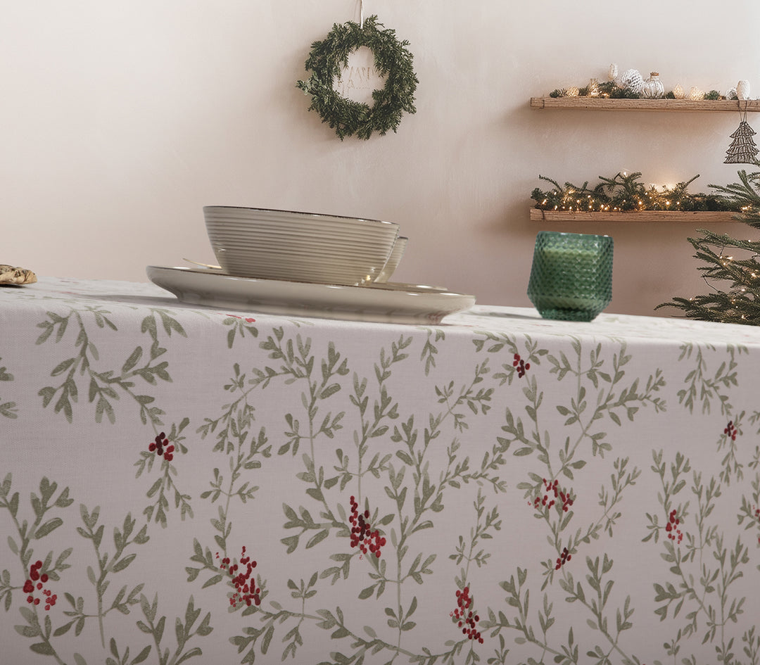 Χριστουγεννιάτικο τραπεζομάντηλο με σχέδιο από πράσινα κλαδιά και κόκκινους καρπούς, στρωμένο σε τραπέζι με πιάτα και πράσινο κερί, σε γιορτινό φόντο με στεφάνι και φωτισμένο δέντρο.