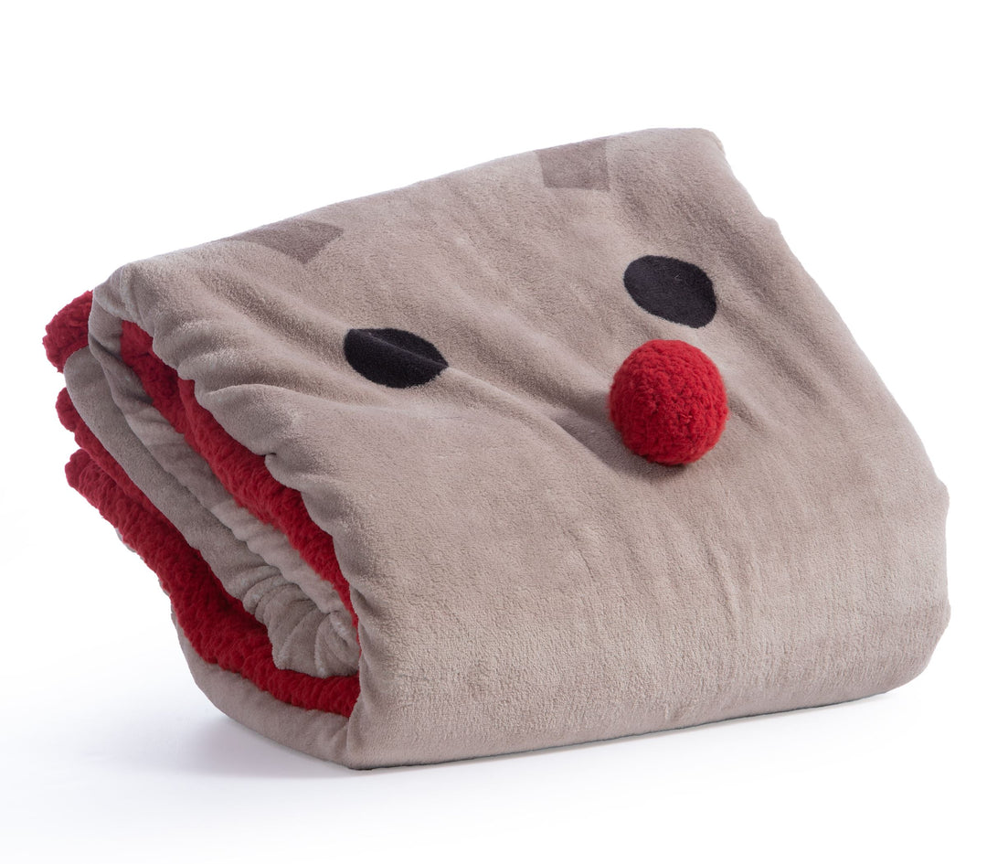 Καφέ fleece κουβέρτα με σχέδιο ταράνδου και κόκκινο περίγραμμα sherpa ριγμένη σε λευκό καναπέ, δίπλα σε ξύλινο τραπέζι με κόκκινες κούπες και χριστουγεννιάτικη διακόσμηση.