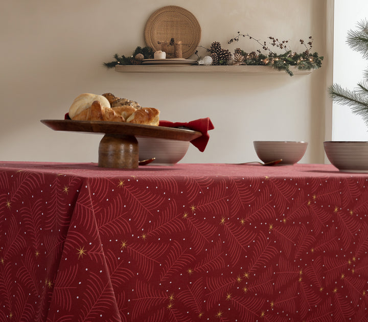 Κόκκινο τραπεζομάντηλο με χρυσά αστέρια και λεπτομέρειες σε σχέδιο κλαδιών, στρωμένο σε ξύλινο τραπέζι με διακόσμηση από φώτα και κεράκια στο φόντο.
