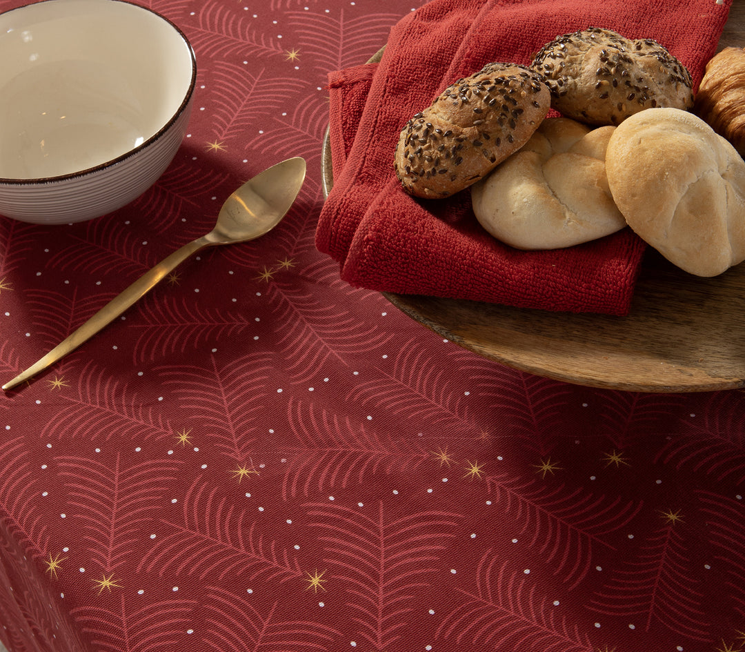 Κοντινό σε κόκκινο τραπεζομάντηλο με χρυσά αστέρια και λεπτομέρειες σε σχέδιο κλαδιών, επάνω του πιατέλα με ψωμάκια και κόκκινες πετσέτες, με κουτάλι και μπολ στο πλάι.