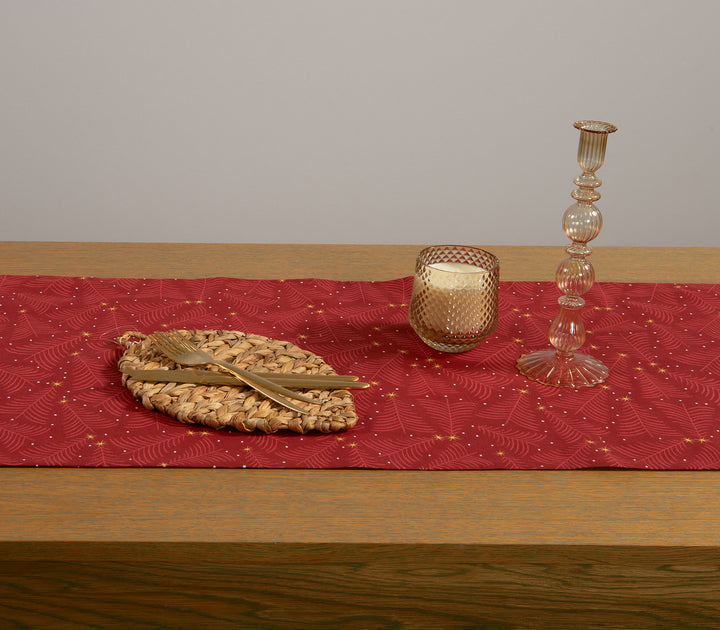Χριστουγεννιάτικος κόκκινος διάδρομος τραπεζιού με μοτίβο χρυσών αστεριών και λεπτομέρειες σε σχέδιο κλαδιών, διακοσμημένος με ψάθινο σουπλά, χρυσά μαχαιροπίρουνα και γυάλινο κηροπήγιο.